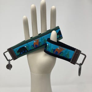 May include: Two keychains with a blue, green, and brown mountain scene with a bear and moose. The keychain on the left is green and the keychain on the right is black.