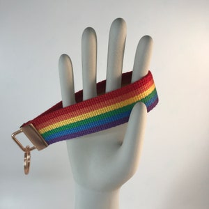 K&ouml;nnte beinhalten: Ein regenbogenfarbener Schl&uuml;sselanh&auml;nger mit einem goldenen Schl&uuml;sselring. Der Schl&uuml;sselanh&auml;nger ist aus gewebtem Stoff mit roten, orangen, gelben, gr&uuml;nen, blauen und violetten Streifen.