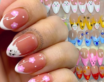 May include: A set of almond-shaped press-on nails featuring a nude base with white French tips. The designs include various cartoon character faces, small pink and yellow flowers, and colorful accents. The text @peachesandmushrooms is visible.