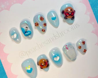 May include: Set of ten blue and white press-on nails with various cartoon designs, including a milk carton, a glass of milk with a straw, a brown bear character, and a heart. The nails are decorated with glitter and rhinestones. The nails are displayed on a white card with the text "@peachesandshrooms" in black.