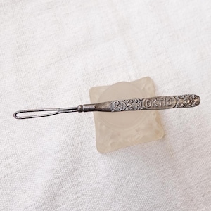 May include: A silver metal threader with an ornate design and the inscription "OKT 13".