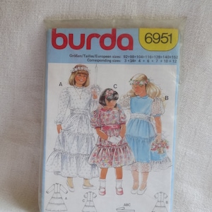 Op de afbeelding: Burda naaipatroon nummer 6951 voor meisjesjurken. Het patroon bevat drie verschillende stijlen: een witte jurk met een geruite rok, een jurk met bloemenprint met een geruite rok en een lichtblauwe jurk met een geruite rok. Het patroon is voor maten 92-152.