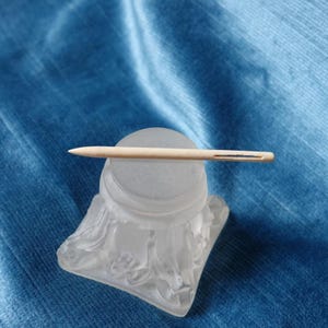 May include: A light-coloured wooden needle resting on a frosted glass object. The needle has a small eye for threading. The glass object has a square base with a decorative, flowing design.