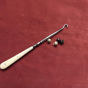 Könnte beinhalten: Ein antiker Häkelhaken mit cremefarbenem Griff und einem Schaft aus Silbermetall. Drei kleine Knöpfe in Beige und Schwarz liegen neben dem Haken. Die Artikel befinden sich auf rotem Hintergrund.