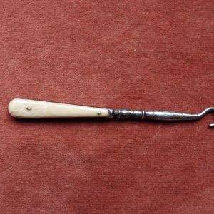 Könnte beinhalten: Ein antikes Schuhhorn mit einem cremefarbenen Griff und einem gebogenen, silberfarbenen Metallhaken. Der Griff ist an der Metallschaft genietet. Das Schuhhorn ist ein Vintage-Artikel, wahrscheinlich zum Anziehen von Schuhen.