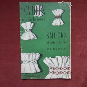以下が含まれることがあります： 緑色の背景に「Les Cahiers du jardin des modes」のタイトルが書かれたヴィンテージ雑誌の表紙。カラフルな刺繍が施された白いスモックのイラストが特徴です。「SMOCKS et points faciles 100 BRODERIES」のテキストも表示されています。