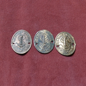 Può includere: Tre medaglioni antichi del marchio Singer Manufacturing Company. Ogni medaglione è di forma ovale con la scritta "THE SINGER MANFG CO" attorno al bordo. Il centro presenta un disegno di macchina da cucire. I medaglioni sono color bronzo e poggiano su una superficie marrone scuro.