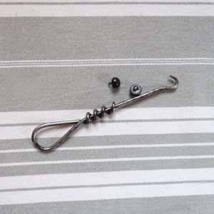 Peut inclure: Un crochet en métal argenté avec une poignée torsadée et un petit bouton noir à l'extrémité. Il y a un deuxième bouton noir à côté du crochet.