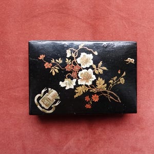 May include: A rectangular black box with a floral design. The box features white and gold flowers with brown and green leaves. A gold emblem is on the lower left corner. The box is on a red surface.