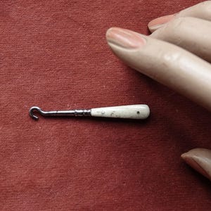 Könnte beinhalten: Ein kleiner, antiker Häkelhaken mit einem Metallschaft und einem weißen Griff. Der Griff hat einen kleinen schwarzen Punkt und ist leicht gebogen. Der Haken liegt auf einer roten Oberfläche.