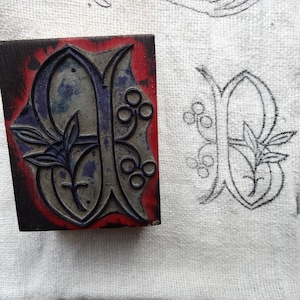 Puede incluir: Un bloque de impresión de madera vintage con un borde rojo y una letra "B" azul y gris con un diseño floral. El bloque se utiliza para crear una letra decorativa para imprimir.