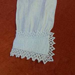 May include: A close-up view of a white fabric sleeve, showcasing detailed lace trim at the cuff. The lace has a scalloped edge and an openwork pattern. The fabric is slightly creased, indicating a soft texture. The cuff is approximately 10 cm wide.