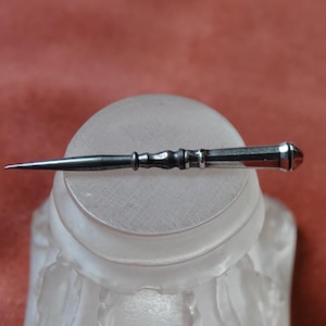 May include: An antique silver needle with a pointed tip and a decorative handle. The needle is lying on a frosted glass surface, with a reddish-brown background. The handle features a small, reddish-brown gem.