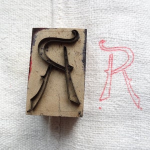 Puede incluir: Un bloque de impresión tipográfica vintage con la letra "R" en una fuente serif decorativa. La letra metálica está montada sobre un bloque de madera rectangular. Una impresión de tinta roja de la letra "R" es visible a la derecha.