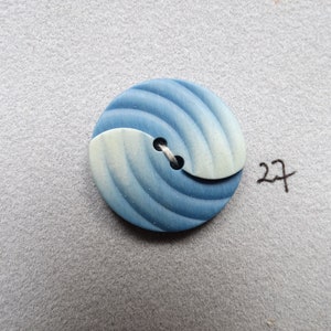 May include: A round button with a blue and white swirl design. The button has a two-tone color scheme, with a light blue and white section and a darker blue section. The button is attached with white thread. The number "27" is written in black ink.