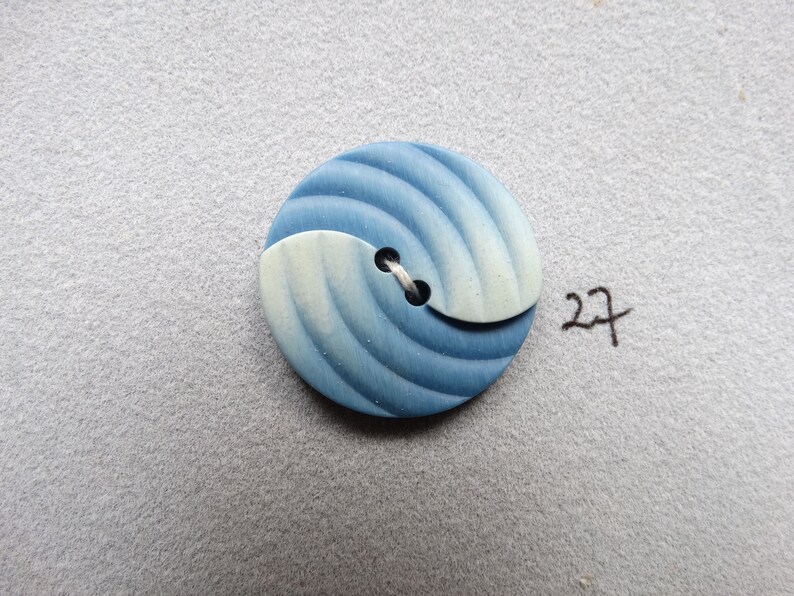 May include: A round button with a blue and white swirl design. The button has a two-tone color scheme, with a light blue and white section and a darker blue section. The button is attached with white thread. The number "27" is written in black ink.