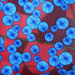 Op de afbeelding: Rode stof met een herhalend patroon van blauwe cirkels met een radiaal ontwerp. De cirkels zijn van verschillende grootte en zijn schijnbaar willekeurig gerangschikt.