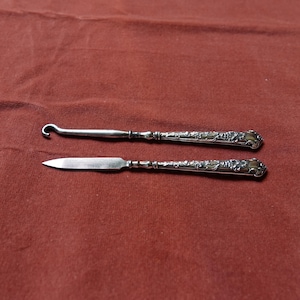 Könnte beinhalten: Zwei antike Silberwerkzeuge, ein Haken und ein Messer, liegen auf einer rotbraunen Oberfläche. Die Griffe sind mit floralen Mustern verziert. Das Hakentool hat ein gebogenes Ende, während das Messer eine spitze Klinge hat. Diese Werkzeuge sind wahrscheinlich zum Nähen oder Sticken.