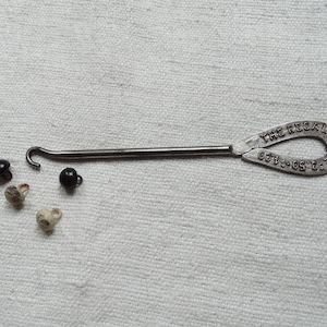 Könnte beinhalten: Ein silberner Knopfhaken mit der Aufschrift "The Regal Shoe Co.". Es befinden sich vier Knöpfe, zwei schwarze und zwei weiße, auf einer weißen Stoffoberfläche.