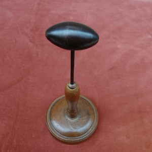 Könnte beinhalten: Ein Vintage-Hutständer mit einer dunkelbraunen, oval geformten Oberseite. Der Ständer hat eine schlanke, dunkle Metallstange, die die Oberseite mit einem gedrechselten Holzsockel verbindet. Der Sockel ist kreisförmig und aus hellbraunem Holz gefertigt.