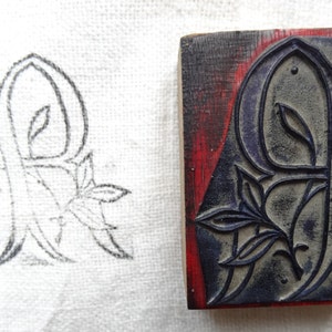Puede incluir: Un bloque de impresión de madera con un diseño tallado de la letra "R" con un motivo floral. El bloque está pintado de rojo y azul.