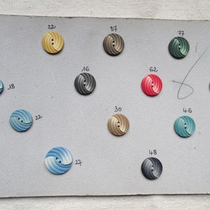 May include: A collection of decorative buttons in various colors, including yellow, gray, red, and blue, arranged on a gray card. Each button features a spiral design and is numbered. The card also has handwritten numbers and text.