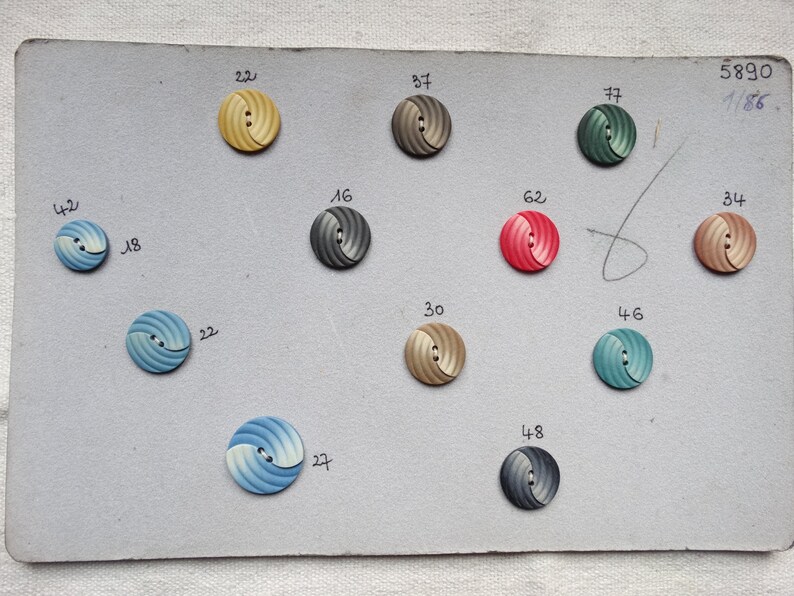 May include: A collection of decorative buttons in various colors, including yellow, gray, red, and blue, arranged on a gray card. Each button features a spiral design and is numbered. The card also has handwritten numbers and text.