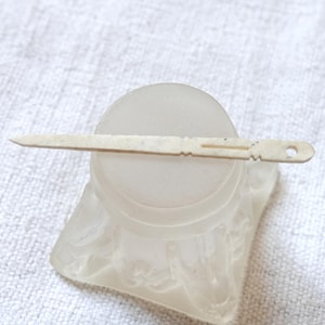 May include: A white bone toothpick with a decorative design and a small hole at the end. The toothpick is resting on a clear glass base.