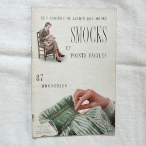 以下が含まれることがあります： 「Smocks et Points Faciles」というタイトルのヴィンテージフランス語の裁縫パターンブック。表紙には87と「Broderies」の文字が書かれています。表紙には刺繍をする人と、座って縫い物をする女性のイラストが描かれています。