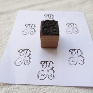 Op de afbeelding: Een houten stempel met een zwart inktkussen met een decoratieve letter B. De stempel ligt op een wit vel papier met meerdere afdrukken van de letter B.