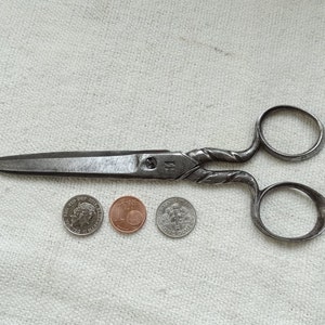 May include: Antique silver-toned scissors with a twisted handle design. The scissors are open and lying on a white fabric surface. Three coins are also on the surface.