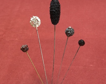 May include: A collection of five hat pins with decorative heads. The pins feature various designs, including a silver filigree, a beaded oval, and carved dark beads. The pins are displayed against a red background.