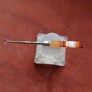 Könnte beinhalten: Ein Vintage-Häkelhaken aus Silber und Achat, mit einem gebogenen Hakenende und einem Griff mit Bändern in Orange, Braun und Weiß. Der Haken liegt auf einem Sockel aus mattiertem Glas vor einem rotbraunen Hintergrund. Ein einzigartiges Werkzeug.