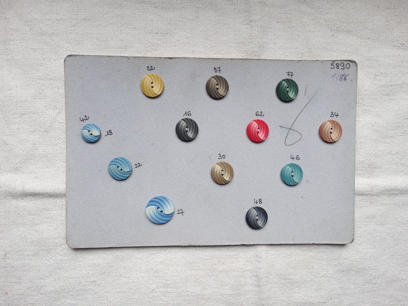 May include: A collection of vintage buttons arranged on a gray card. The buttons are in various colors, including yellow, blue, gray, red, and brown. Each button has a spiral design and is labeled with a number. The card has the numbers "5890" and "1186" written on it.