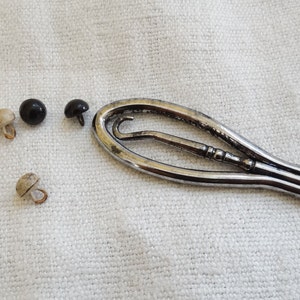 Può includere: Uno strumento per bottoni in argento con un gancio a un'estremità e un piccolo strumento appuntito all'altra. Ci sono quattro bottoni su una superficie di tessuto bianco: due bottoni neri e due bottoni beige.