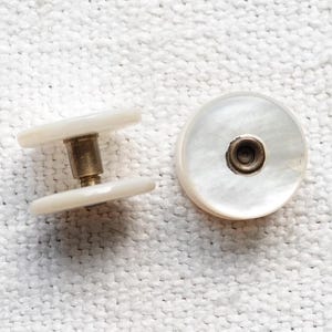 以下が含まれることがあります： 真珠母貝の白いヴィンテージボタン2つと真鍮製の金具。1つのボタンは組み立てられており、2つの真珠ディスクの間の真鍮製のポストを示しています。もう1つのボタンは、真鍮の中心部を持つ単一の真珠ディスクです。