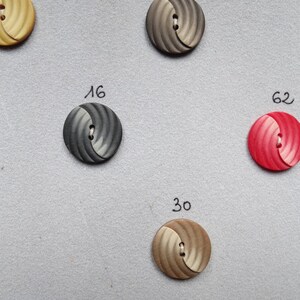 May include: A collection of round buttons in various colors, including beige, gray, and red. Each button has a spiral design and a two-hole center. Numbers are written next to some of the buttons, such as "16" and "62".