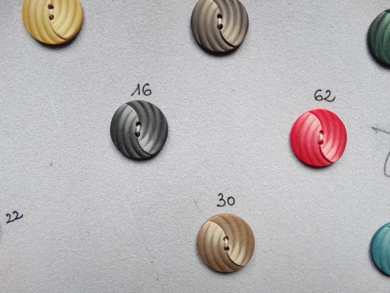 May include: A collection of round buttons in various colors, including beige, gray, and red. Each button has a spiral design and a two-hole center. Numbers are written next to some of the buttons, such as "16" and "62".