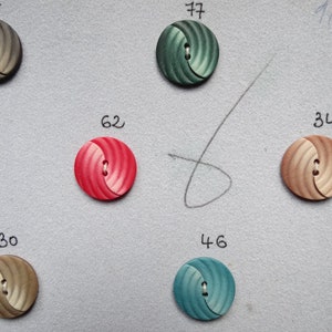 May include: Assortment of decorative buttons in various colors, including gray, green, red, and tan. Each button has a spiral design and a two-hole center. Numbers are written above each button.