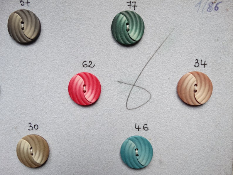 May include: Assortment of decorative buttons in various colors, including gray, green, red, and tan. Each button has a spiral design and a two-hole center. Numbers are written above each button.