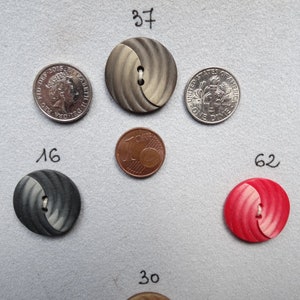 May include: Assortment of decorative buttons in various colors and designs, including gray, red, and green. The buttons are arranged on a gray surface, with a U.S. dime and a quarter for size comparison. The buttons have two or four holes for sewing.