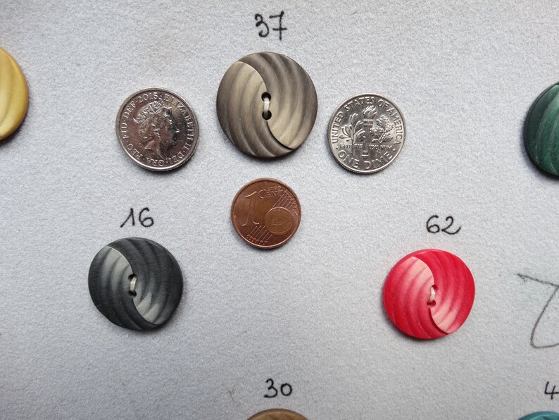 May include: Assortment of decorative buttons in various colors and designs, including gray, red, and green. The buttons are arranged on a gray surface, with a U.S. dime and a quarter for size comparison. The buttons have two or four holes for sewing.