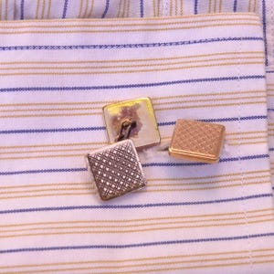 Op de afbeelding: Close-up van drie vierkante manchetknopen in goud- en zilverkleuren. De manchetknopen bevinden zich op een gestreept wit en blauw overhemd. Twee manchetknopen zijn goudkleurig en één is zilverkleurig met een patroon.