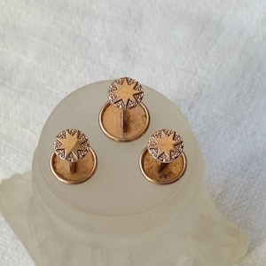 Puede incluir: Tres gemelos dorados con un diseño de estrella en el centro. Los gemelos están sobre una superficie blanca.