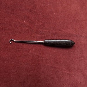 May include: A metal hook tool with a dark wooden handle. The hook is silver and curved, and the handle is a dark brown color. The tool is lying on a red surface.
