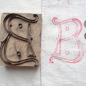 Puede incluir: Un bloque de impresión de madera con un diseño de letra "B" tallada. El bloque se utiliza para crear una impresión de tinta roja de la letra en una superficie de tela blanca. También se pueden ver tres monedas de plata en la tela.