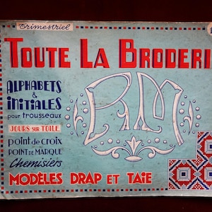 May include: A vintage French embroidery pattern book titled "Toute La Broderie." The cover is light blue with red and white text, featuring the title, "Alphabets & Initiales," and other embroidery-related terms. Decorative elements include a stylized "RM" and a corner pattern.