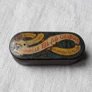 Puede incluir: Una caja de hojalata vintage con un diseño azul y dorado. El texto en la caja dice "Pour coudre Solidement Employez le Fil au Chinois en pelotes au Fil d'Or".