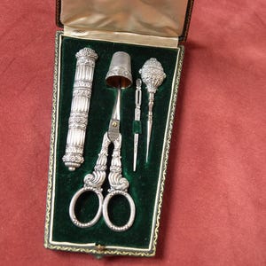 Könnte beinhalten: Ein silbernes Nähset in einem grün mit Samt ausgekleideten Etui. Das Set enthält eine verzierte Schere, einen Fingerhut, ein Nadelgehäuse und zwei weitere Werkzeuge. Das Etui ist geöffnet und zeigt den Inhalt vor rotem Hintergrund.