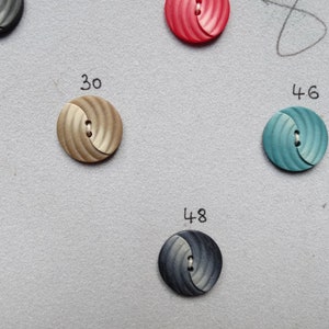 May include: Close-up of several round buttons in various colors, including tan, teal, and navy. Each button has a swirled design and two holes for sewing. Numbers 27, 30, 46, and 48 are written near the buttons.